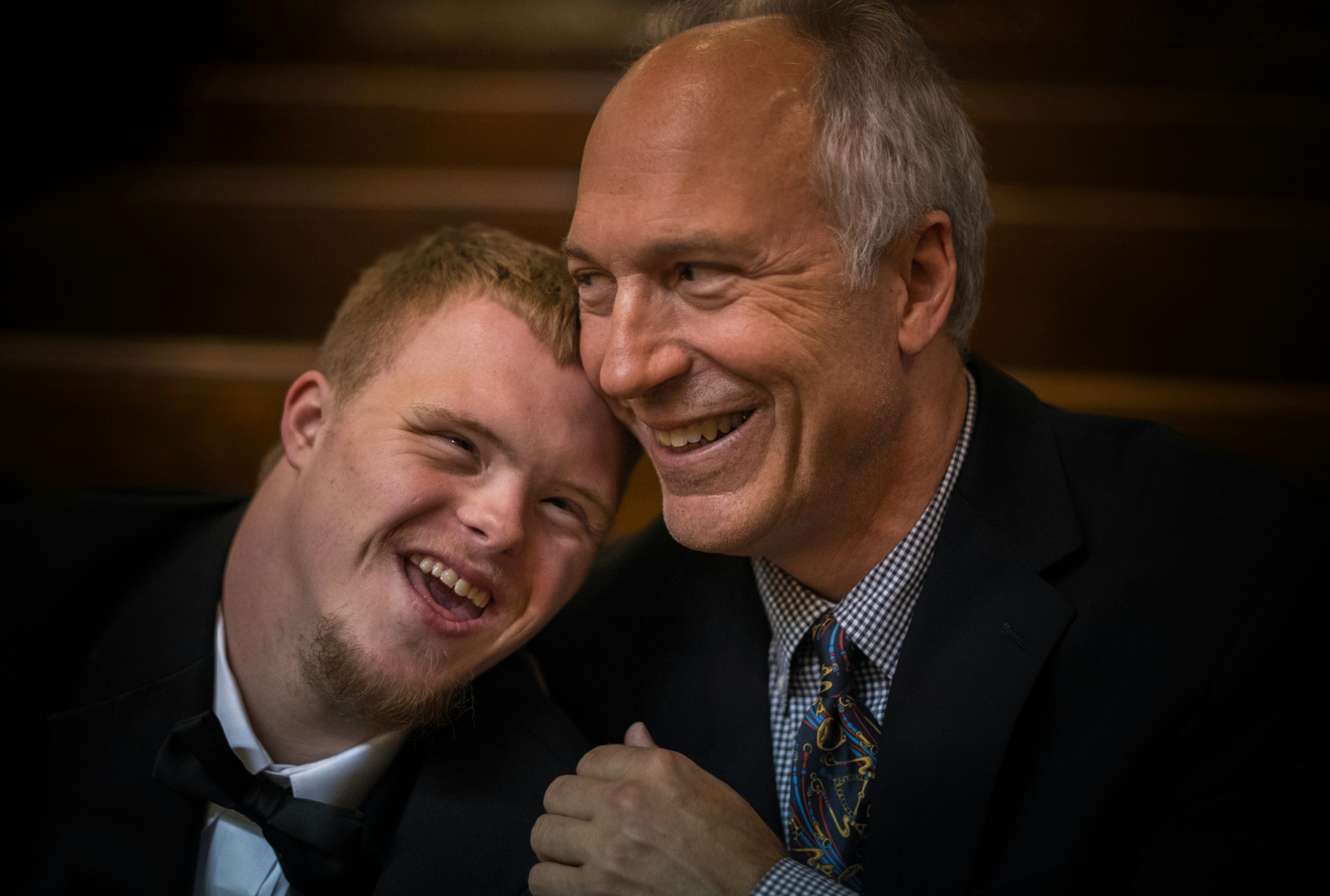 Two men smiling and enjoying a moment together, one with red hair and a beard wearing a tuxedo, the other with gray hair wearing a suit and tie, in a warm indoor setting.